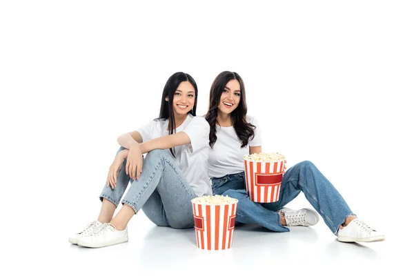 Brunette interracial women smiling at camera while sitting near big buckets of popcorn on white — Fotografia de Stock