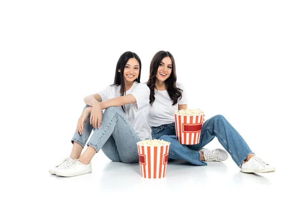 Brunette interracial femmes souriant à la caméra tandis que assis près de gros seaux de maïs soufflé sur blanc — Photo de stock