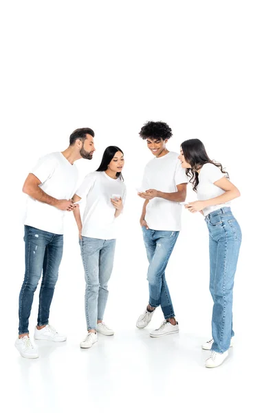 Vista completa de sonriente afroamericano hombre chateando en el teléfono inteligente cerca de curiosos amigos interracial en blanco - foto de stock