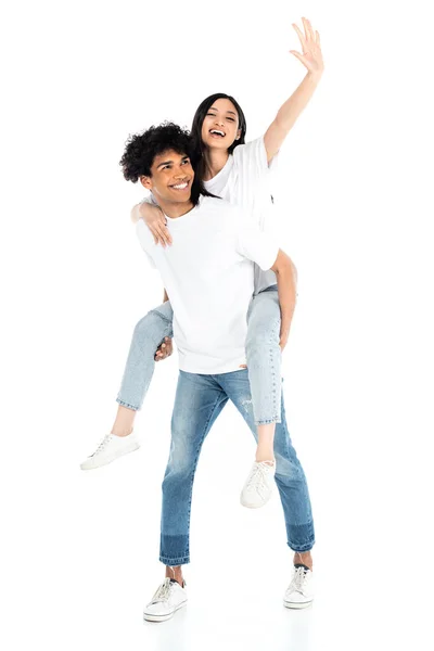 Smiling african american man piggybacking excited asian woman waving hand on white — Stockfoto