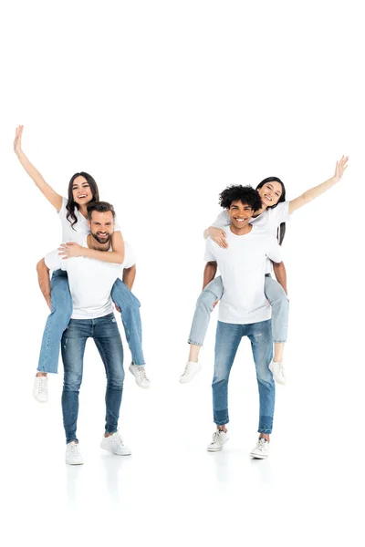 Full length view of interracial men piggybacking cheerful women waving hands on white — Stock Photo