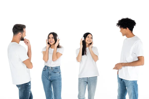 Hombres interracial mirando alegres mujeres jóvenes escuchando música en auriculares aislados en blanco - foto de stock