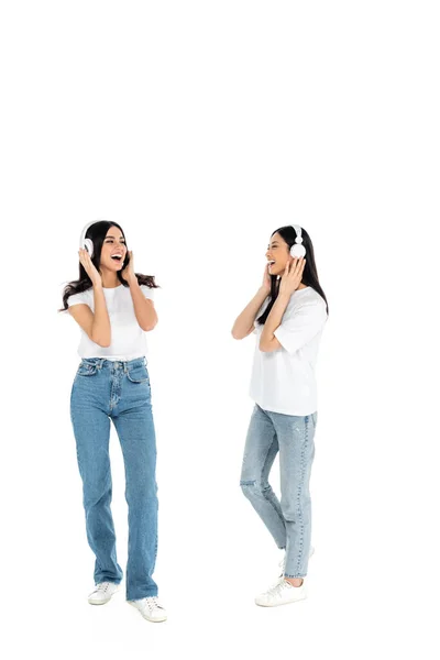 Full length view of excited multiethnic women in jeans and headphones listening music on white - foto de stock