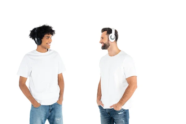 Interracial men in headphones standing with hands in pockets of jeans and looking at each other isolated on white - foto de stock