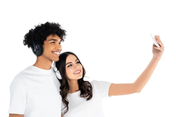 Cheerful interracial couple in headphones taking selfie on mobile phone isolated on white — Stock Photo