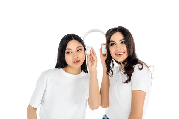 Happy interracial women holding headphones and listening music isolated on white - foto de stock