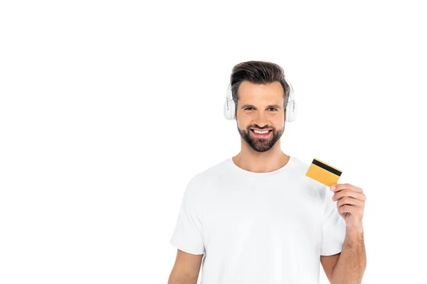 Hombre complacido en los auriculares mirando a la cámara mientras sostiene la tarjeta de crédito aislada en blanco - foto de stock