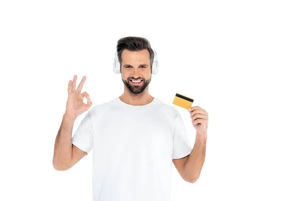 Hombre complacido en auriculares con tarjeta de crédito y mostrando gesto bien aislado en blanco - foto de stock