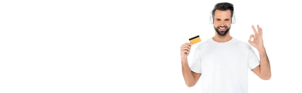 Homem sorrindo em fones de ouvido segurando cartão de crédito e mostrando gesto ok isolado em branco, banner — Fotografia de Stock