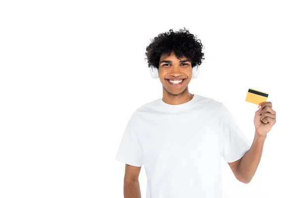 Young african american man in headphones holding credit card and smiling at camera isolated on white — Stockfoto