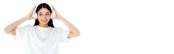 Alegre asiático mujer tocando auriculares mientras escucha música aislado en blanco, bandera - foto de stock