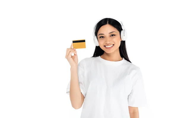 Усміхнена азіатська жінка в навушниках тримає кредитну картку ізольовано на білому — стокове фото