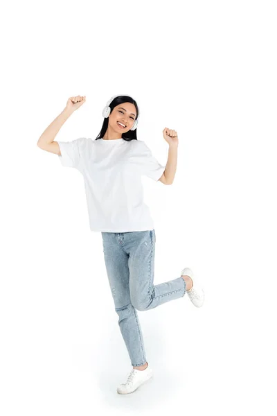 Full length view of pleased asian woman in jeans and headphones dancing on white — Fotografia de Stock