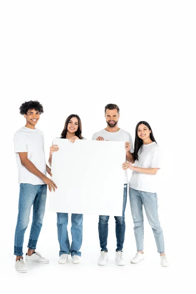 Vue pleine longueur des amis multiethniques heureux en jeans tenant une pancarte vierge sur blanc — Photo de stock