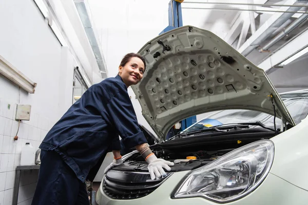 Smiling mechanic in uniform standing near car with open hood — Stockfoto