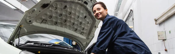 Mecánico positivo mirando lejos cerca del coche con capucha abierta en el garaje, pancarta - foto de stock