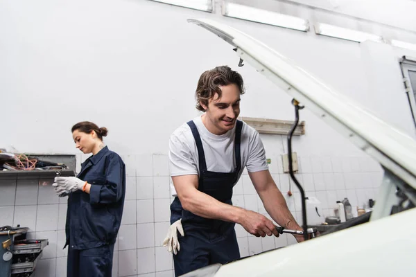 Mécanicien positif travaillant avec une clé et une voiture près d'un collègue flou en service — Photo de stock