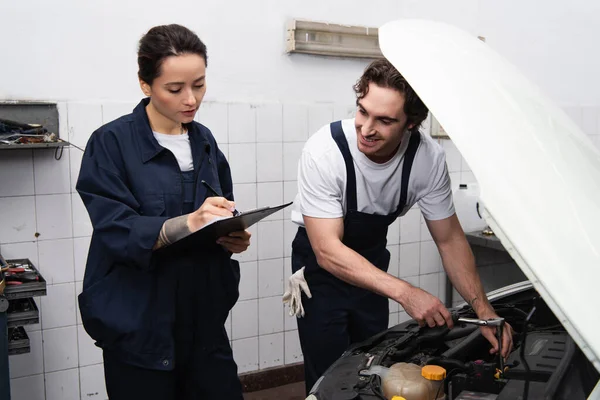 Mechanic writing on clipboard near smiling colleague and car in garage — Stock Photo