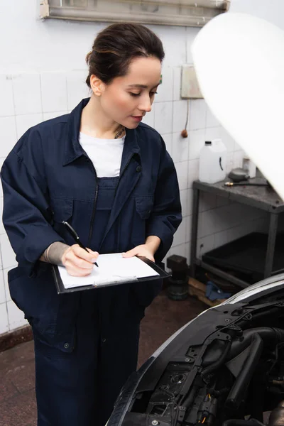 Giovane meccanico in uniforme che scrive sugli appunti e guarda l'auto in servizio — Foto stock