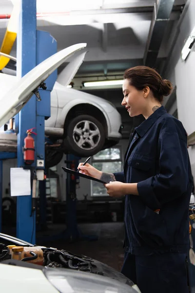 Side view of tattooed mechanic writing on clipboard near car in service — Stock Photo