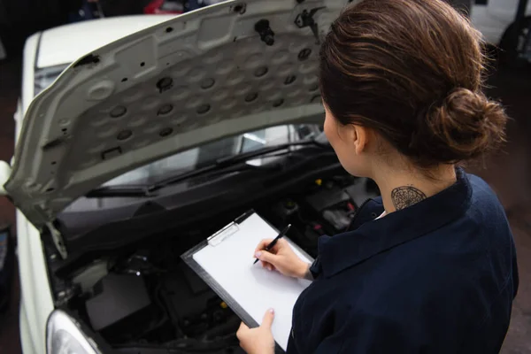 High angle view of mechanic writing on clipboard near car with open hood in service — стоковое фото