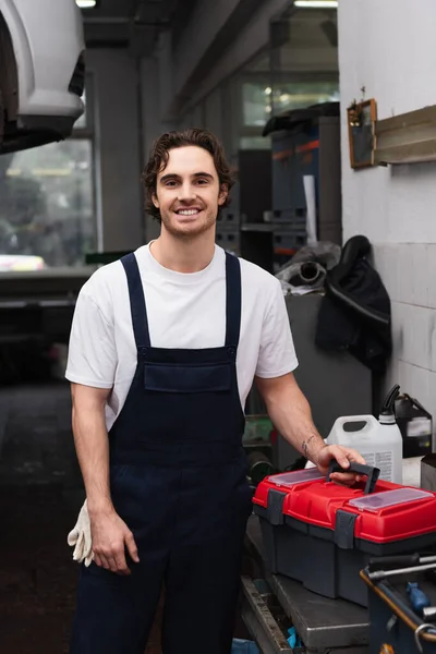 Cheerful mechanic in overalls looking at camera near toolbox in garage — Stock Photo