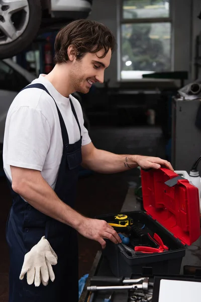 Vista lateral del trabajador sonriente mirando las herramientas en la caja de herramientas en el servicio de automóvil - foto de stock