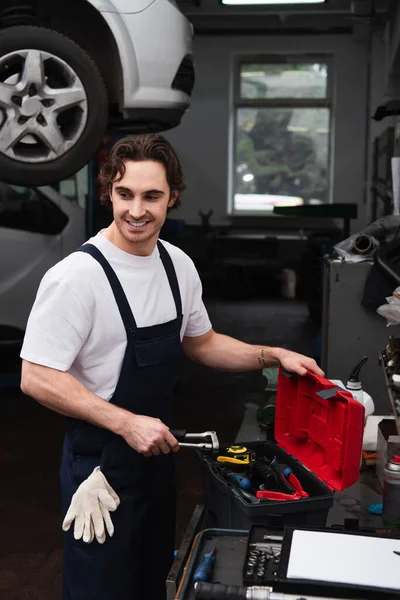 Mecánico sonriente de pie cerca de herramientas y caja de herramientas en servicio de automóviles - foto de stock