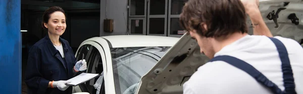 Reparaturfrau in Handschuhen hält Klemmbrett neben verschwommenem Kollegen und Auto in Garage, Banner — Stockfoto
