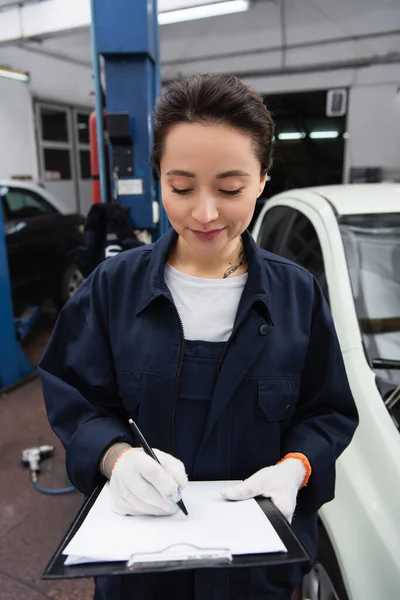 Tattooed workwoman writing on clipboard near car in service — Stockfoto