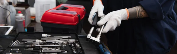 Cropped view of tattooed mechanic holding tool near toolbox in car service, banner — стоковое фото