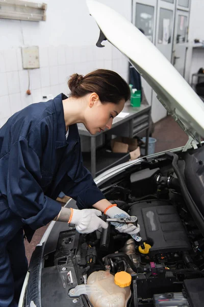 Side view of workwoman holding wrench near auto with open hood in garage — Stock Photo