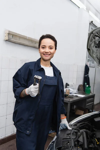 Positive mechanic holding tool and looking at camera near car in garage — Stockfoto