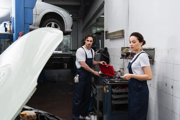 Mecânica em uniforme olhando para o carro com capuz aberto em serviço — Fotografia de Stock