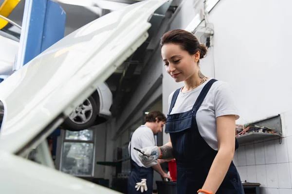 Positive mechanic holding wrench near car with blurred open hood in service — Stock Photo