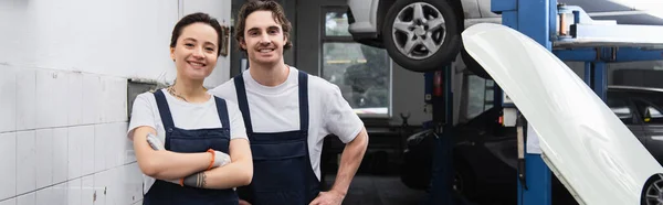 Young mechanics smiling at camera near hood of car in service, banner — Stock Photo