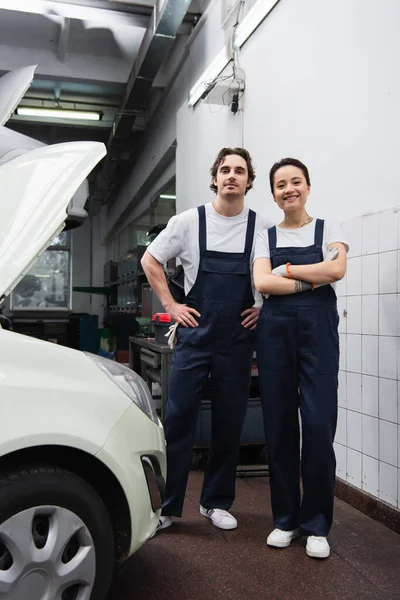 Young mechanics in overalls looking at camera near car with open hood in garage — Stock Photo