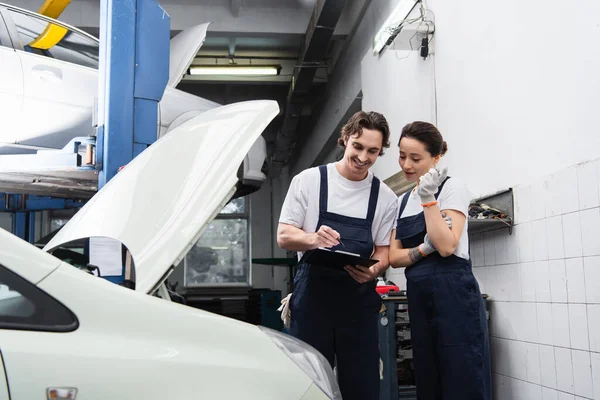 Mechanics with clipboard talking near car with open hood in garage — Stockfoto