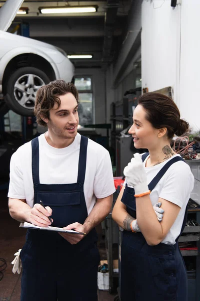 Meccanico sorridente in uniforme e guanti che parla con collega con appunti in servizio auto — Foto stock