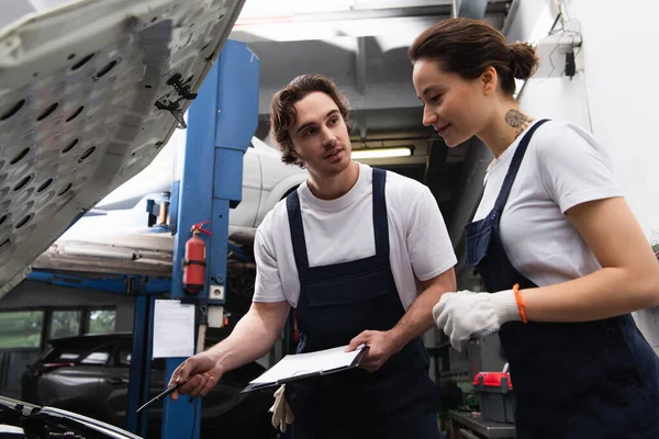 Mechanic with clipboard pointing at car with open hood near colleague in garage — Stock Photo