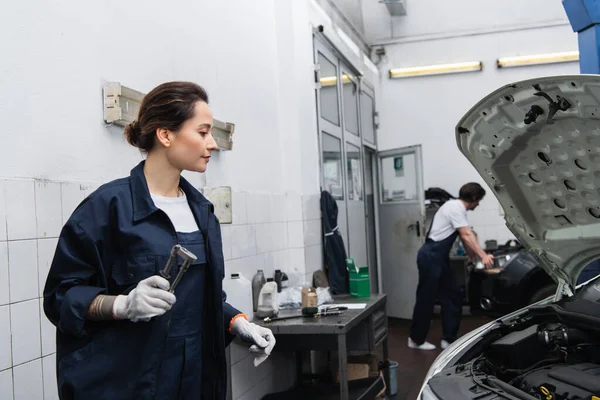 Side view of workwoman in gloves holding tool near auto with open hood in garage — Fotografia de Stock