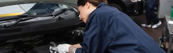 Young workwoman holding tool near car with open hood in service, banner — Stock Photo