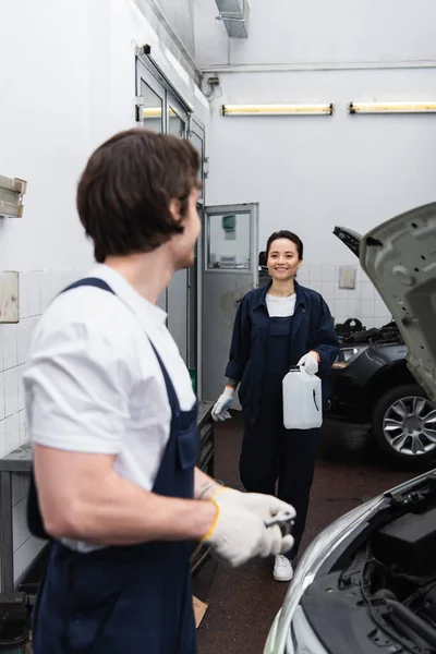 Lächelnder Mechaniker hält Kanister neben verschwommenem Kollegen und Dienstwagen — Stockfoto
