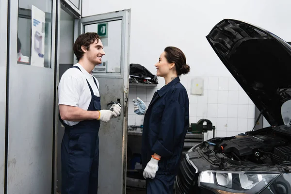 Vista lateral do mecânico sorrindo falando perto do carro com capuz aberto na garagem — Fotografia de Stock