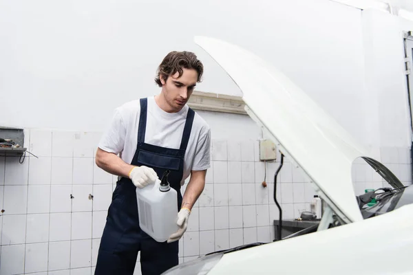 Trabalhador segurando recipiente perto de carro com capuz aberto na garagem — Fotografia de Stock