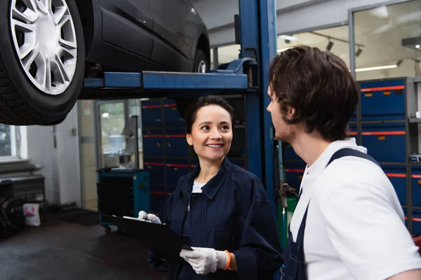 Smiling mechanic writing on clipboard and talking to colleague in car service — Stock Photo