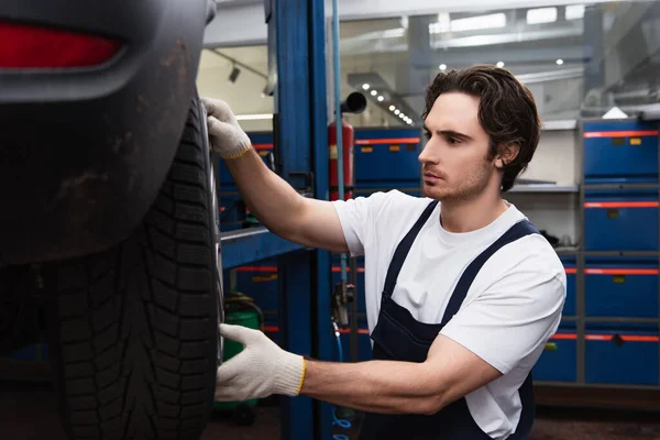 Joven mecánico trabajando con disco de rueda y coche en garaje - foto de stock