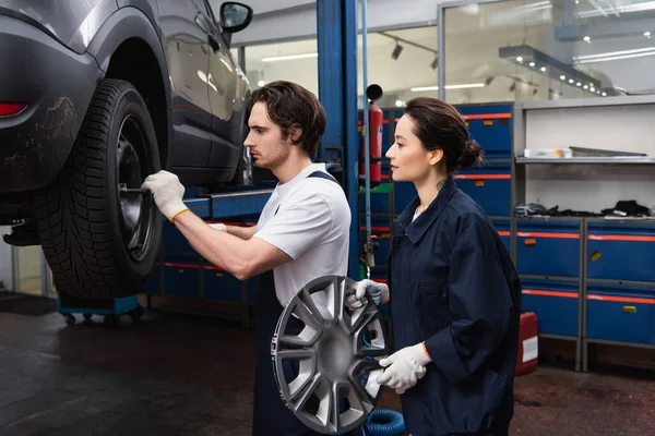 Side view of workwoman holding wheel disk near colleague and auto in garage — Stock Photo