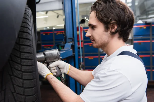 Side view of mechanic working with impact wrench on car wheel in garage — Stock Photo