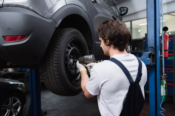 Mecânico em luvas e macacões usando chave de impacto enquanto trabalhava com roda de carro em serviço — Fotografia de Stock